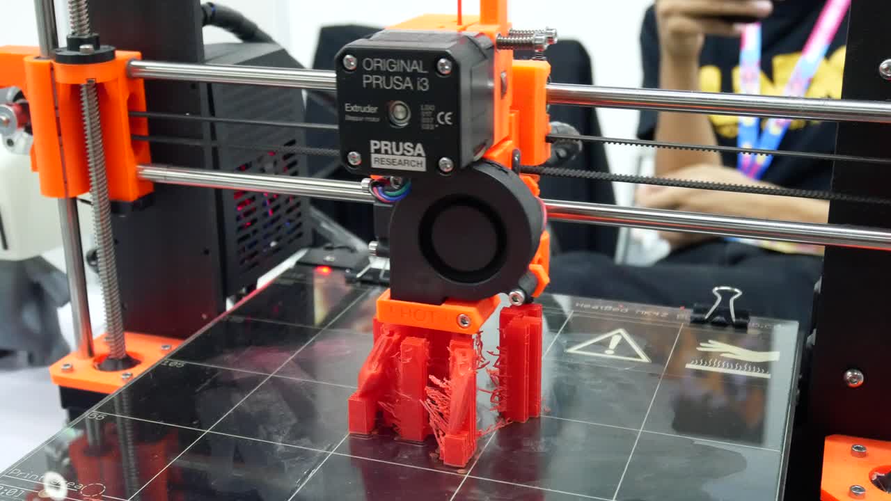 A close-up view of an Original Prusa i3 3D printer in operation, fabricating a red plastic object.