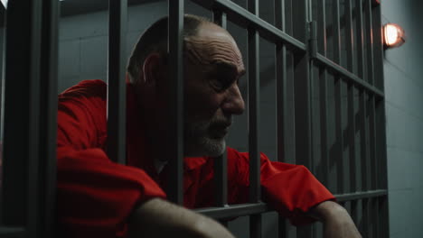 elderly prisoner in orange uniform holds hands on metal bars