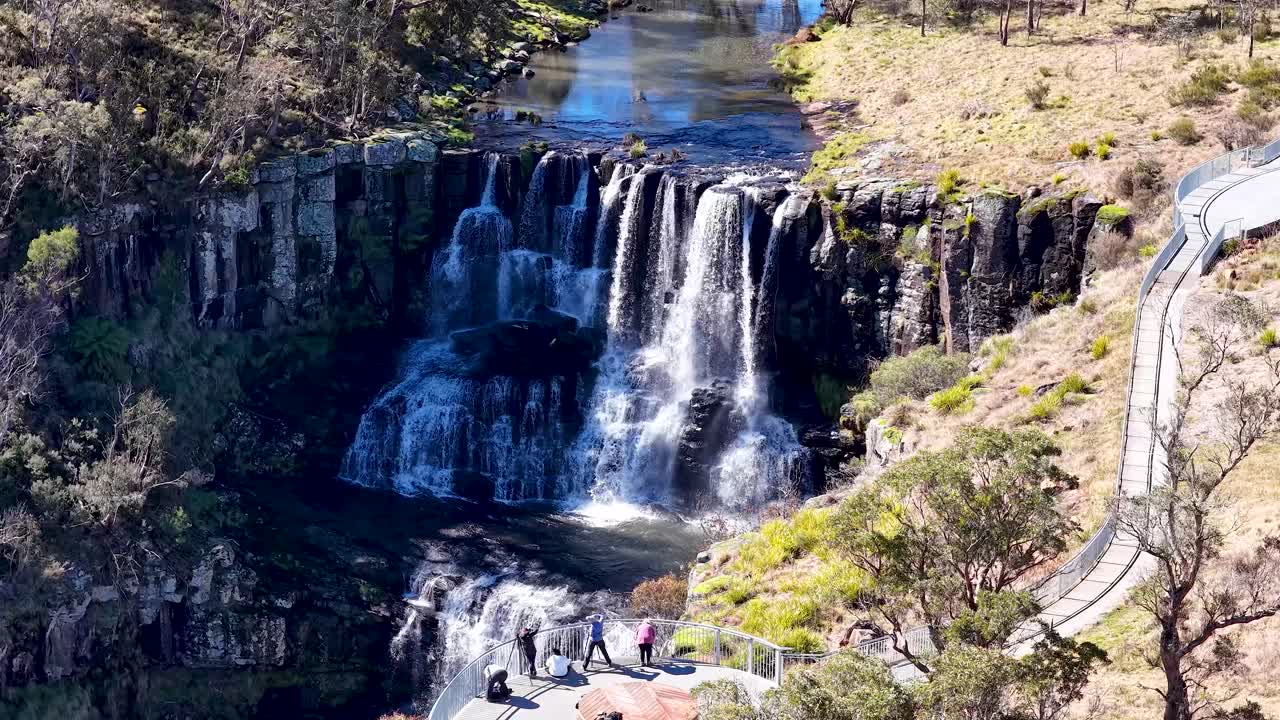 High-angle aerial footage captures Ebor Falls cascading over cliffs, with visitors on a curved walkway under bright daylight in a rugged Australian landscape
