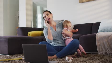 Caucasian-mother-holding-her-baby-talking-on-smartphone-while-working-from-home