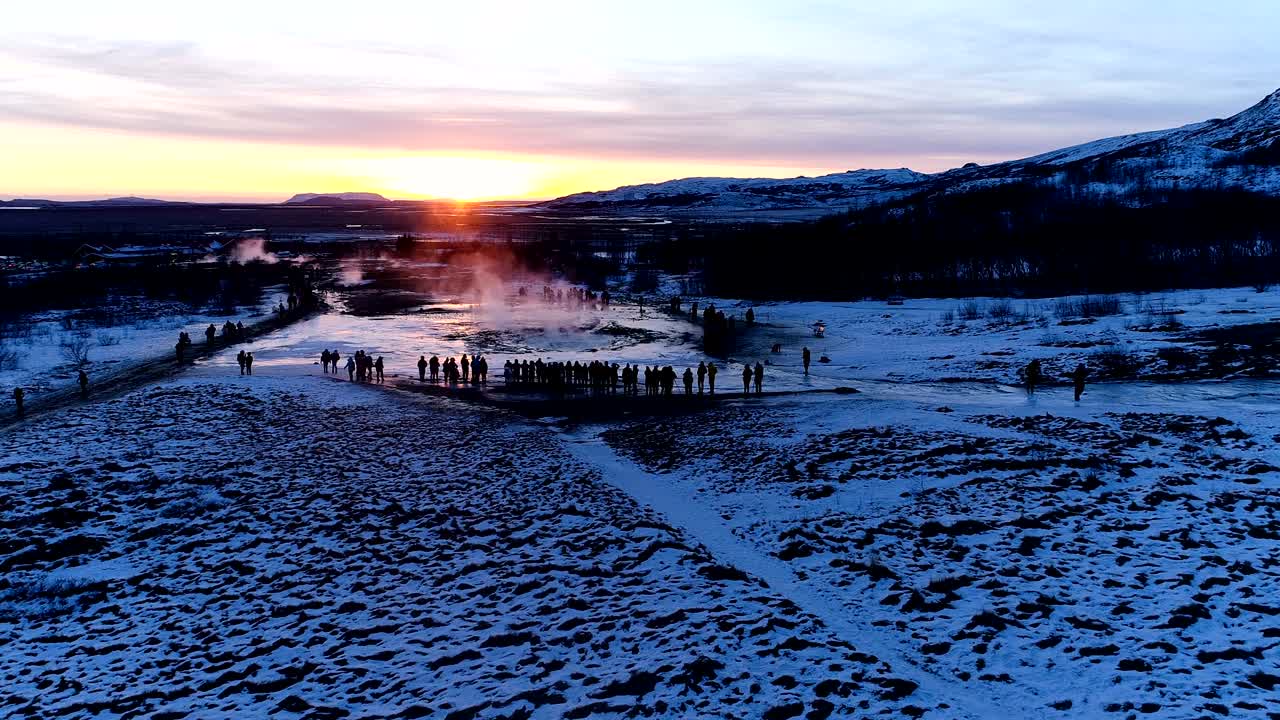 아이슬란드 게이시르의 게이저의 파노라마 뷰: 빨간색과 오렌지색의 해가 지는 에 사람들이 게이저를 둘러싸고 고있는 게이저에서 연기가 나오고 있습니다.
