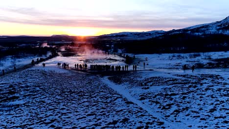 vista panorámica de un geyser en geysir, islandia. el humo está saliendo del geyser con la gente caminando alrededor del geyser, bajo la puesta de sol roja y naranja.