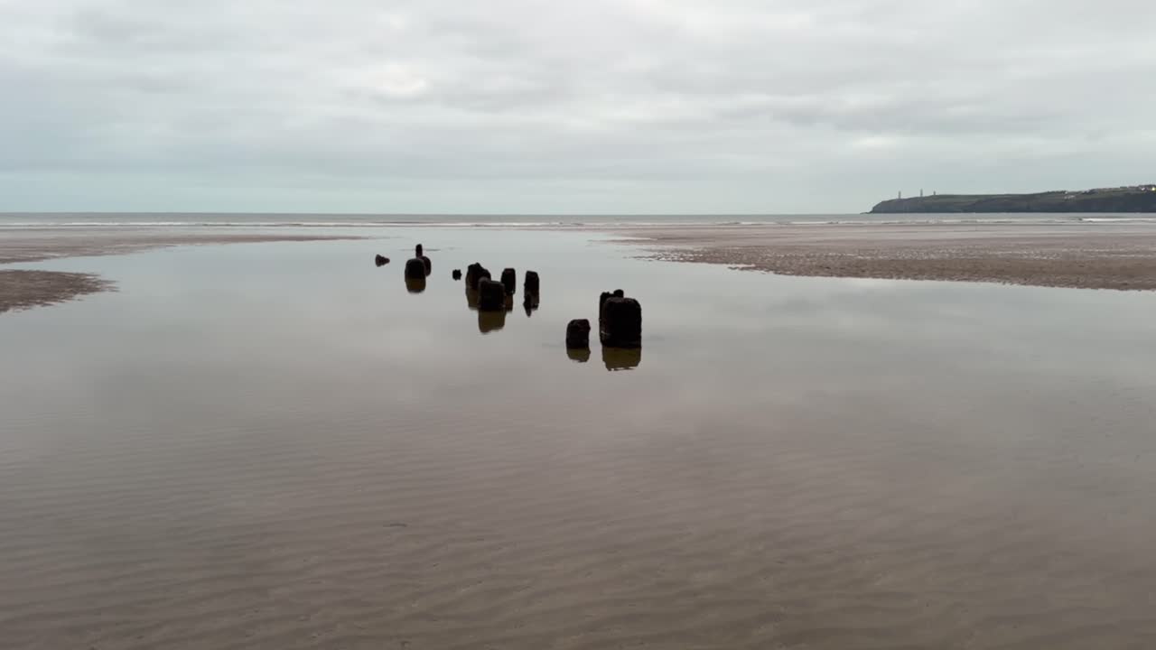 Rotten pilings of old wooden wharf poke out of beach sand, overcast