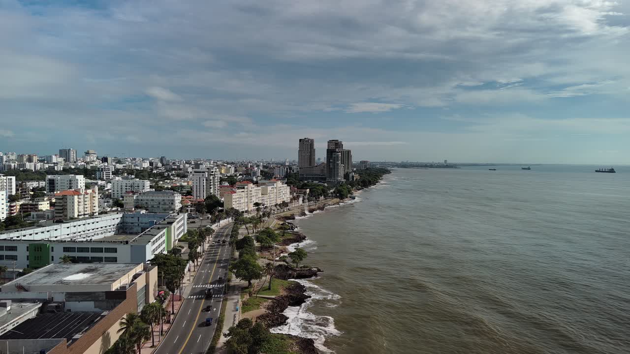 Aerial drone view of the Malecón coastline in Santo Domingo. Features the Caribbean Sea, George Washington Avenue, and the modern city skyline of the Dominican Republic capital
