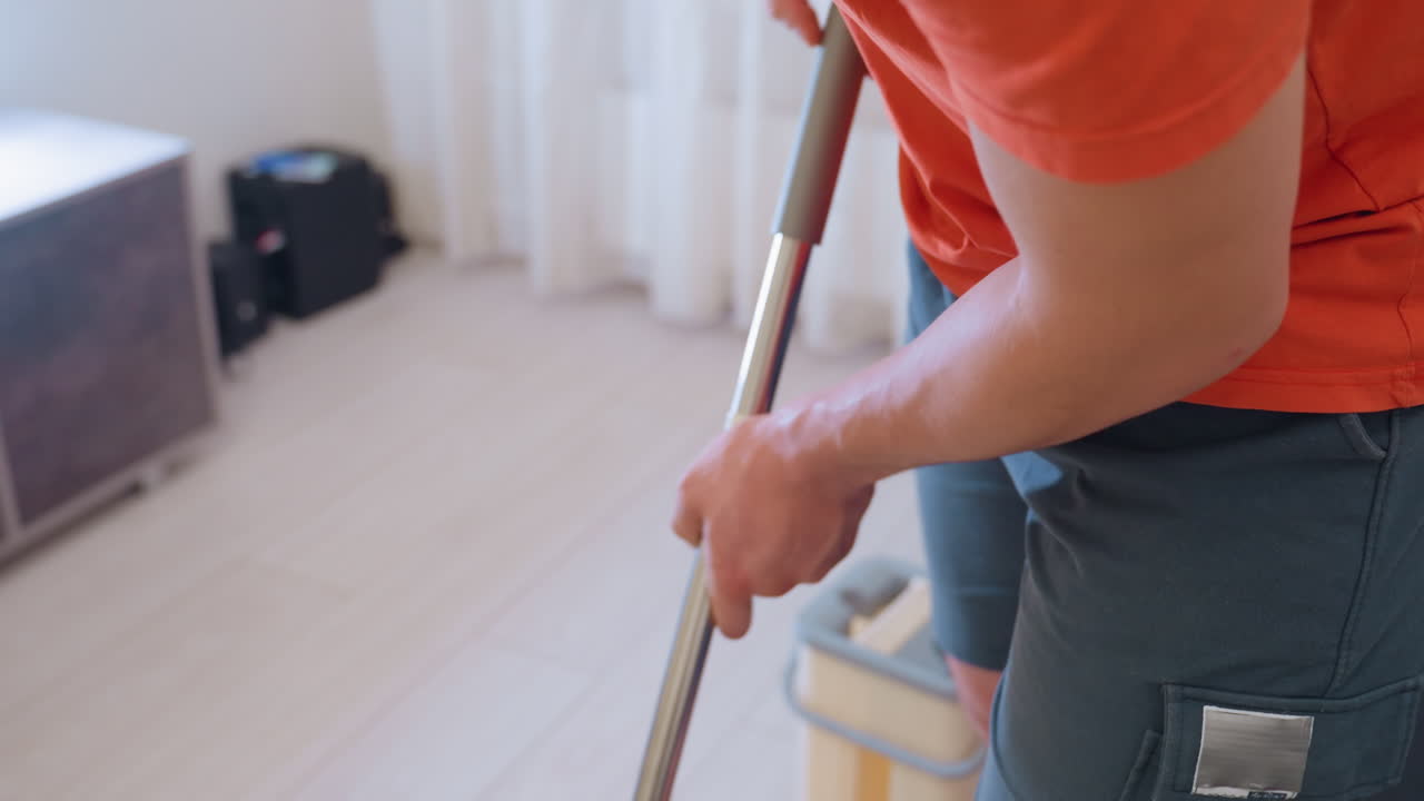 Close up of man in orange shirt and gray shorts gripping mop handle while cleaning floor indoors showing focus on household hygiene cleanliness and daily domestic routine