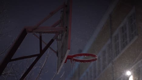 snow falls around basketball backboard and rim with no net at night