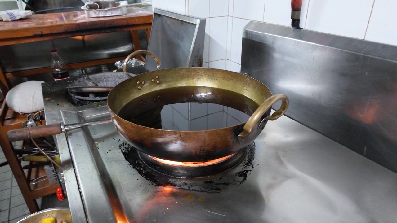 Static medium shot of oil heating in a brass wok over a gas burner flame.