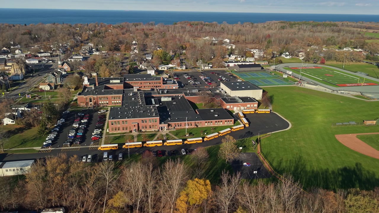 Vista aerea con drone di un campus scolastico negli Stati Uniti con una lunga fila di scuolabus gialli, campi sportivi e aree di parcheggio durante il periodo del rientro a scuola