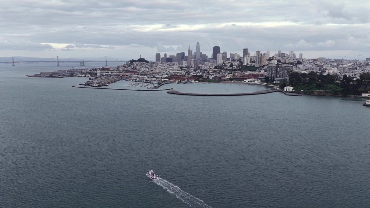 Båt skjærer hvit kjølvann over San Francisco Bay under sentrums skyline, California, USA, innrammet av Bay Bridge, marina, åsside-nabolag under grå overskyet himmel. Bredt luftfoto med drone