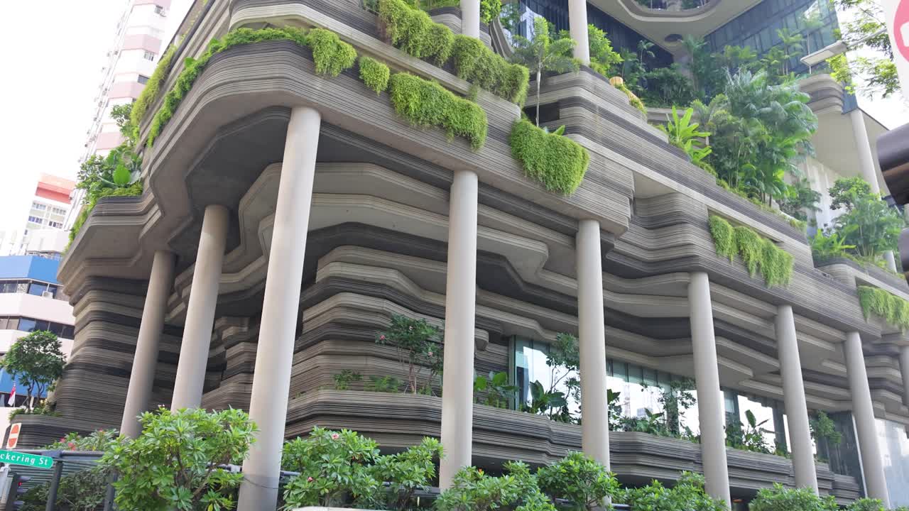 Vehicles pass a multi-story building with lush greenery, filmed in daylight with upward pan