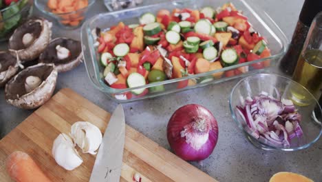 Chopped vegetables in baking dish, chopping board and ingredients on kitchen worktop, slow motion