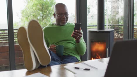 Un-Hombre-Mayor-Afroamericano-Sonriente-Relajándose-Con-Los-Pies-En-Alto,-Usando-Un-Teléfono-Inteligente-Y-Tomando-Café