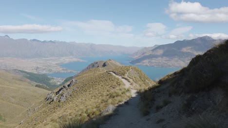 Mountain-path-leading-down-from-Ben-Lomond-with-Lake-Wakatipu,-mountains-and-Queenstown-behind-on-a-sunny-summer-day-in-New-Zealand