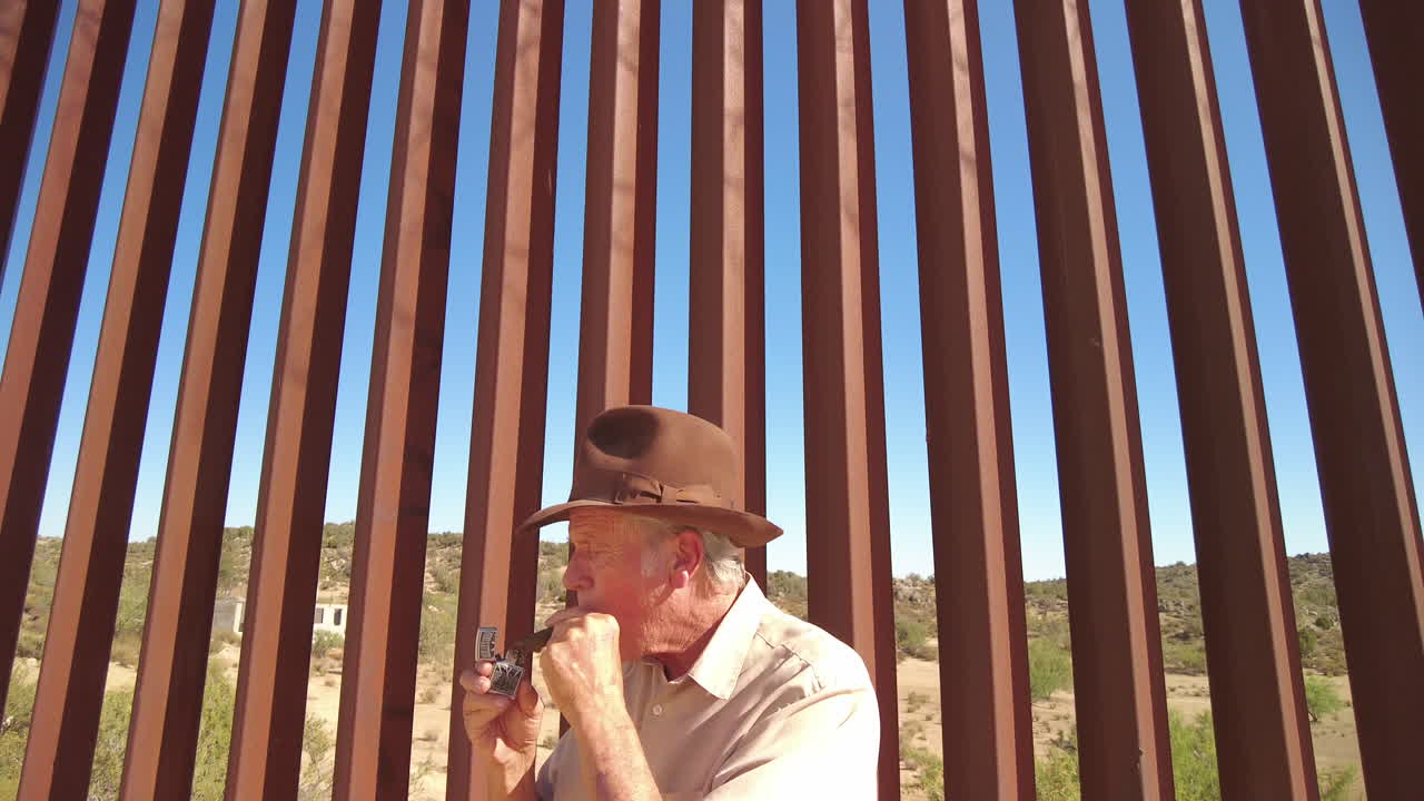 American man lights cigar in front of US Mexico border fence, tilt up to reveal barbed wire