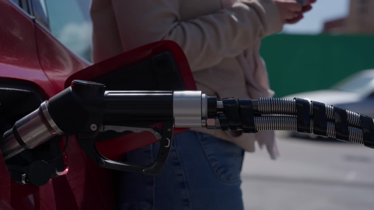 Wanita Mengisi Bahan Bakar Mobil Merah di SPBU Sambil Menggunakan Ponsel