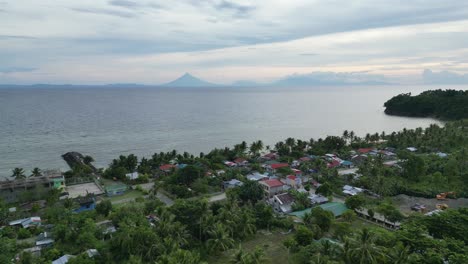 Aerial-View-of-Rural-Beachfront-Town-with-silhouette-of-Mayon-Volcano-looming-in-background