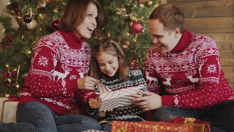 Happy-Little-Girl-Unwrapping-Gift-While-Sitting-With-Her-Mom-And-Dad-In-Front-Of-Christmas-Tree-At-Home-1