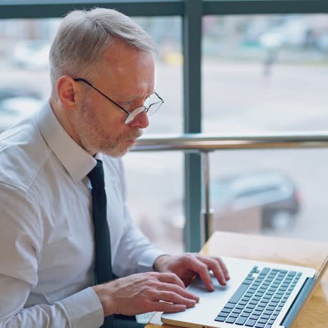 freelance job. mature man working on a laptop near the window. serious businessman typing on a computer indoors on city background.