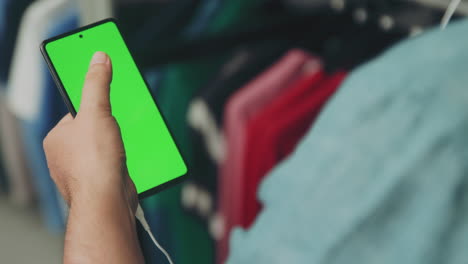 Man-in-Shirt-Using-Mobile-Phone-with-Green-Screen-Scrolling-through-the-Display-while-Watching-and-Listening-through-the-Headphones-in-a-Clothes-Store