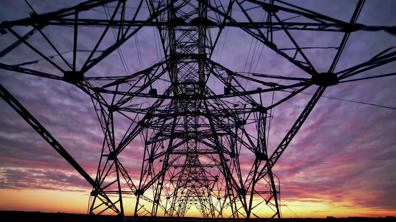 Dramatic low-angle shot of a power line tower against a vibrant sunset sky, capturing the industrial
