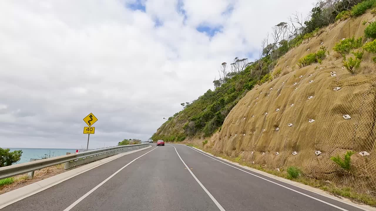 A car travels along the winding Great Ocean Road, surrounded by lush greenery and ocean views under a partly cloudy sky