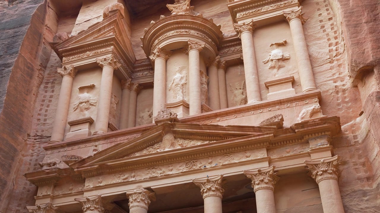 Dettagli intricati in arenaria dell'iconico Al Khazneh a Petra, in Giordania, che mostrano colonne nabatee ornate, intagli e una monumentale facciata antica, ripresa fluida con panoramica verso l'alto a mano libera