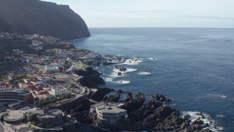 Wide-view-of-serene-coastal-city-by-vast-ocean,-waves-rolling-in,-volcanic-rocks-in-the-water,-natural-pools-and-epic-mountain-in-the-background