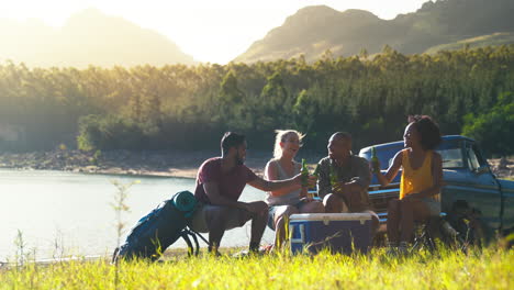 group of friends with backpacks by pick up truck on road trip drinking beer from cooler by lake