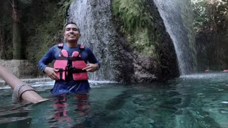 Hombre-Hispano-Hablando-Usando-Un-Chaleco-Salvavidas-En-Un-Río-Azul-Con-Cascada-En-El-Fondo