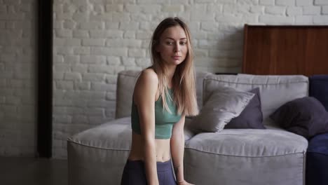 blonde girl unfolding fitness mat on floor at home, looking to the camera