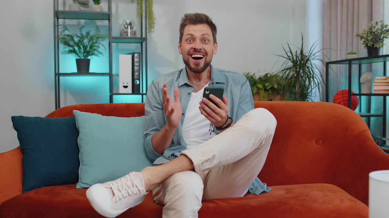 HOMBRE FELIZ Y APUESTO EN EL SOFÁ DE CASA USANDO EL SMARTPHONE, ESCRIBIENDO, NAVEGANDO, EXCLAMANDO 'WOW', 'SÍ', AL DESCUBRIR UNA GRAN VICTORIA