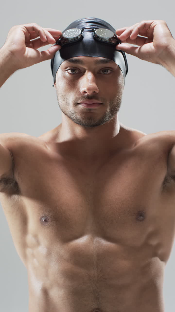 Vertical video: Biracial male athlete, swimmer, adjusting swimming goggles, gray background