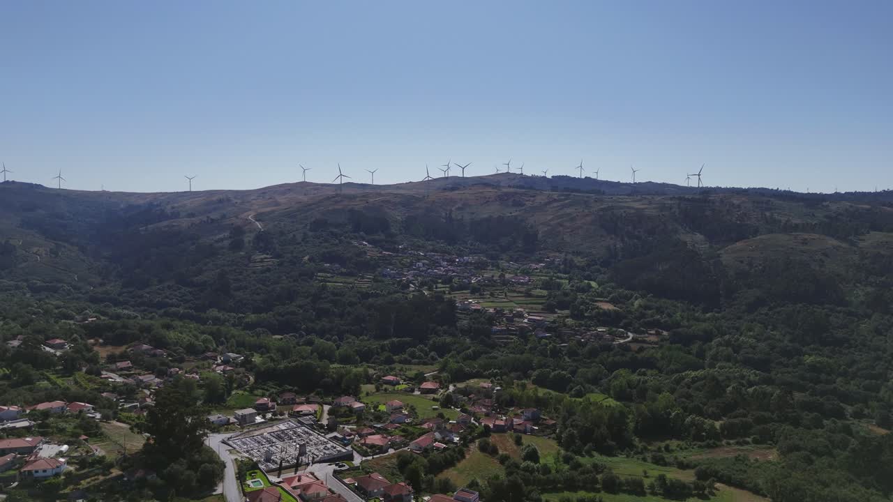 vista aérea de una aldea rural enclavada entre verdes colinas, turbinas eólicas salpican el paisaje lejano