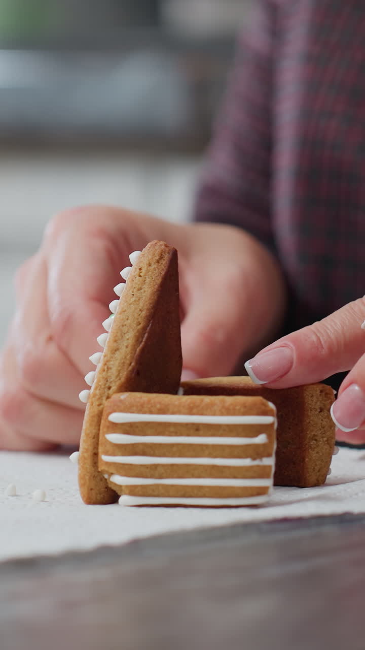 Primo piano di una donna che usa biscotti di pan di zenzero per progettare una casetta, circondata da biscotti a forma di fiore, sac à poche e altri elementi essenziali per la cottura su un tavolo da cucina