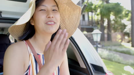 happy asian woman with sun hat sitting in car trunk and blowing kiss on sunny day, slow motion