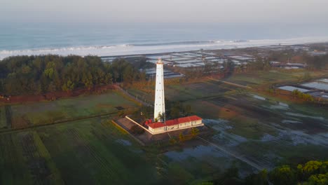 aerial view of white lighthouse surrounded by plantation with beach on the background - ketawang beach, indonesia