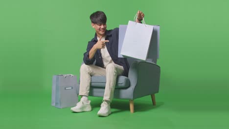 un hombre de compras asiático señalando bolsas de compras antes de sonreír a la cámara mientras está sentado en el sofá frente a la pantalla verde