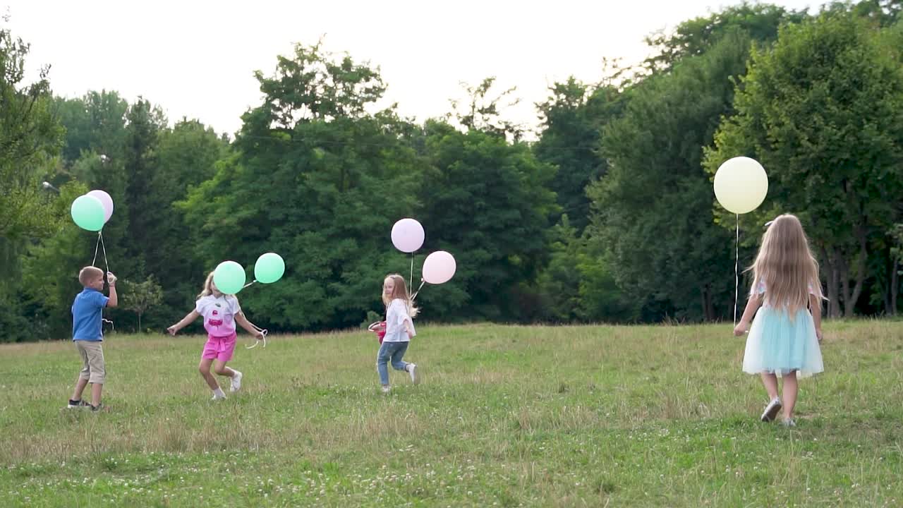 Happy children run on the grass in the park. Boy and girls with balloons in their hands. Summer rest. Children's holiday. Slow motion
