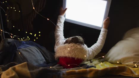 niño caucásico con sombrero de santa, acostado en almohadas en el suelo y usando tableta