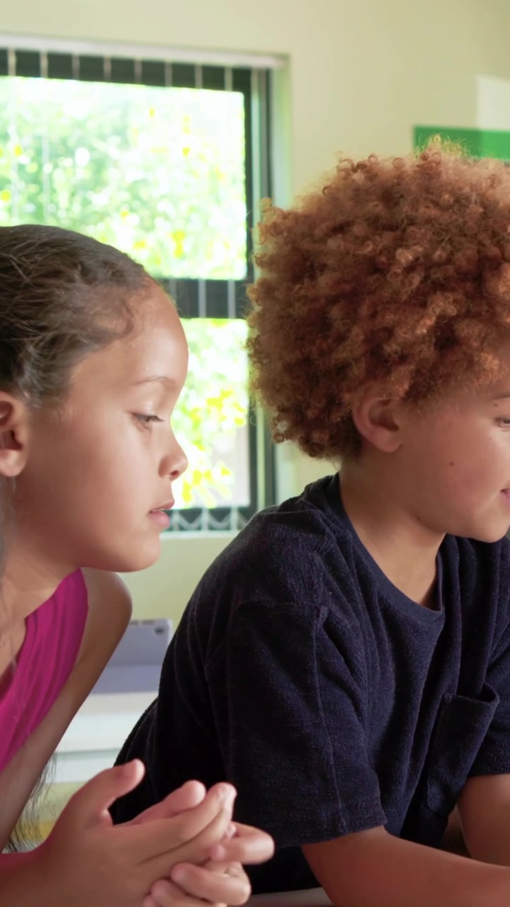 Vertical video: Moving closer magenta-top girl leaning at classroom desk, helping peer on tablet