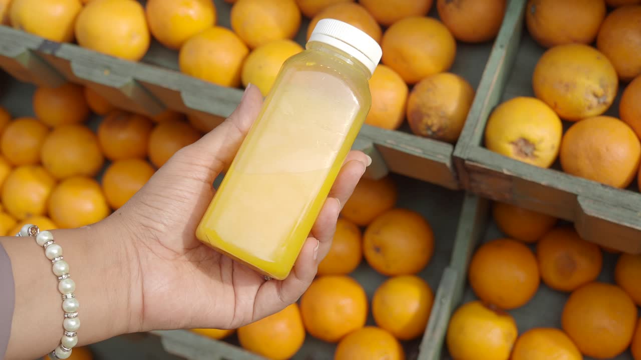 persona sosteniendo una botella de jugo de naranja fresco frente a una caja de naranjas