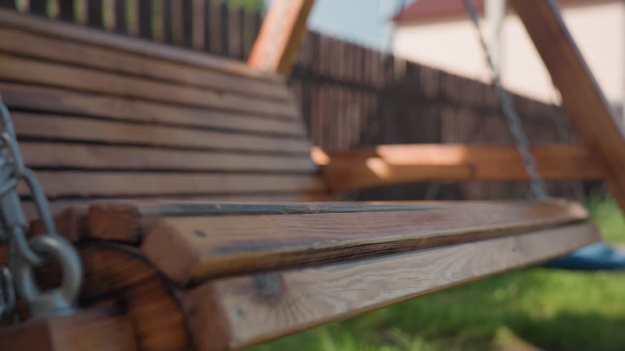 Close up of smooth wooden bench swing gently swaying on metal chains in sunny backyard, with grass below, blurred modern house in distance, and playground style blue swing visible to one side