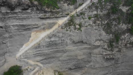 tourists going down the stairway carved into a limestone cliff, aerial dolly out shot revealing secluded diamond beach, nusa penida, bali, indonesia