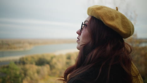 lady in yellow beret and sunglasses wearing a black coat, focusing upward, with a blurred background featuring a river and autumn trees