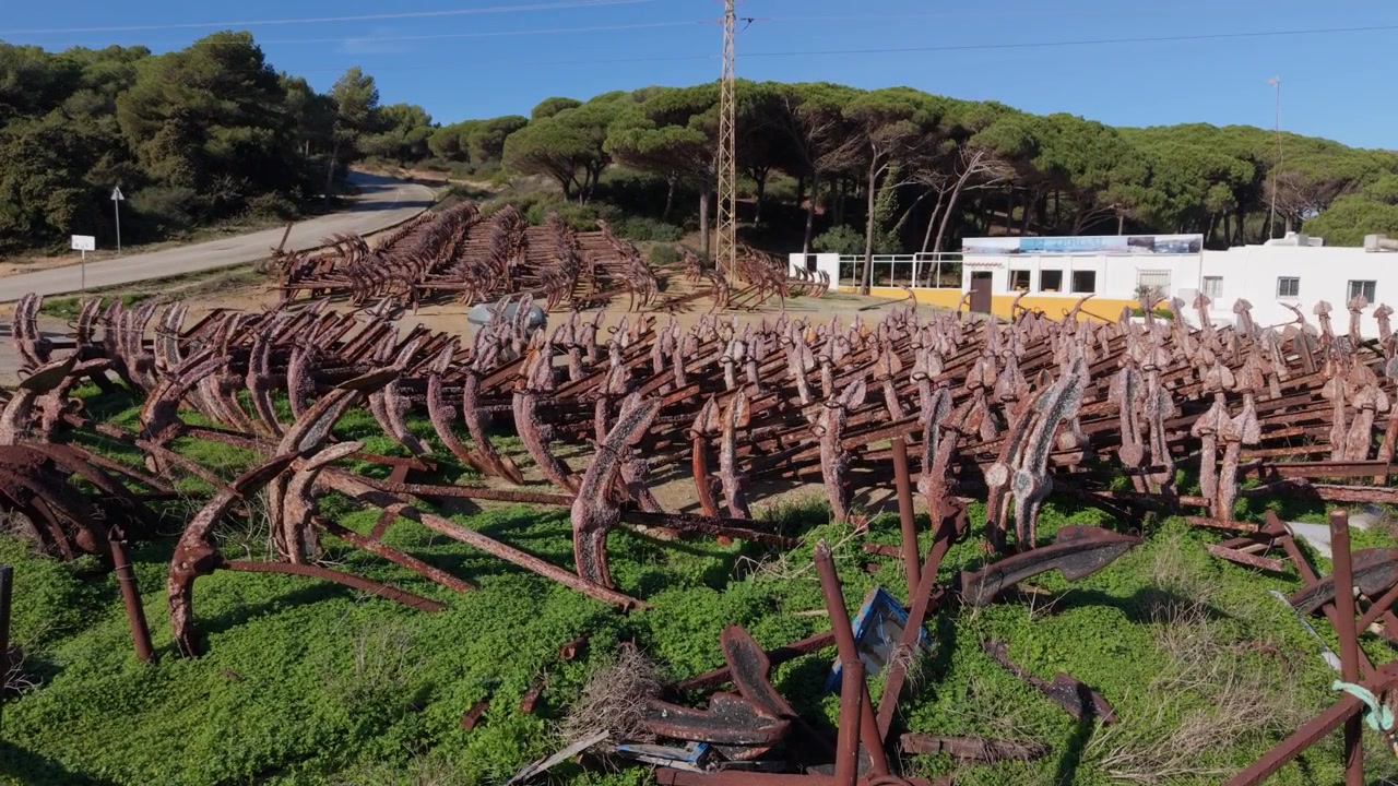 Porto di Conil, Cadice, Spagna. Riprese aeree con drone a bassa quota di centinaia di vecchie ancore immagazzinate lungo il porto con lo sfondo di una pineta sotto un sole splendente con cielo azzurro limpido
