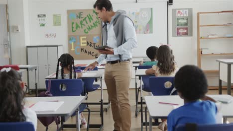 Diverse-male-teacher-with-tablet-helping-children-at-desks-in-elementary-school-class,-slow-motion