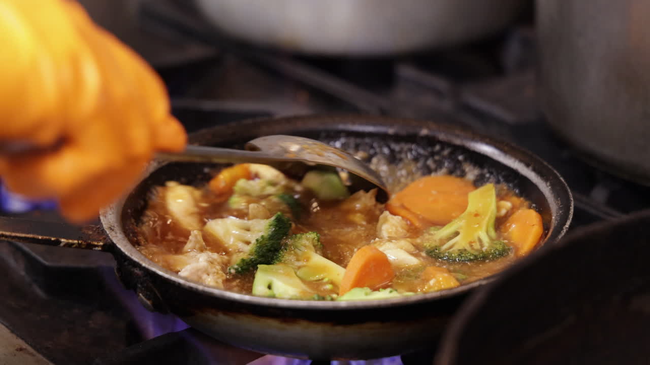 갈색 소스로 끓인 야채 치킨을 요리하고, 요리사는 레스토랑 주방에서 슬롯형 스푼으로 중국식 볶음 요리를 고열로 저어주고, 슬로우 모션 슬라이더는 4k를 닫습니다.