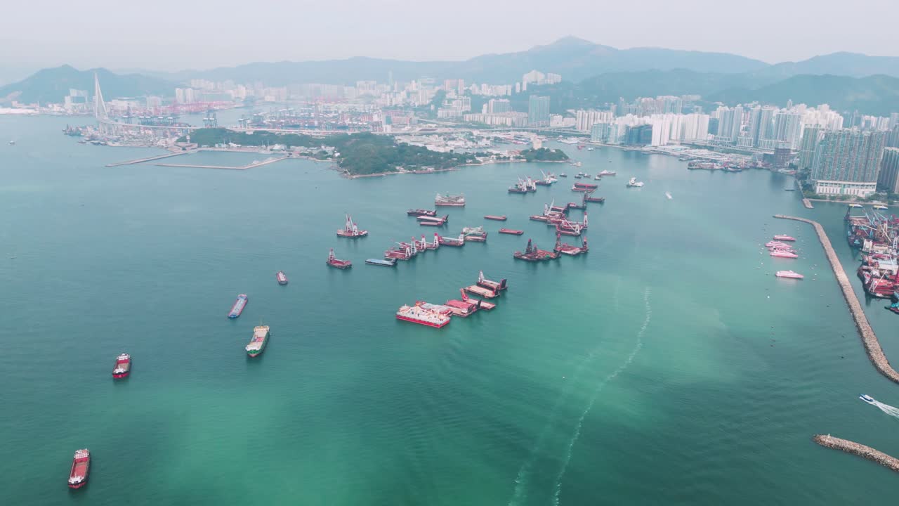 Veduta aerea panoramica del Porto di Hong Kong in una giornata di sole, con navi da carico, terminal container e lo skyline urbano circostante.