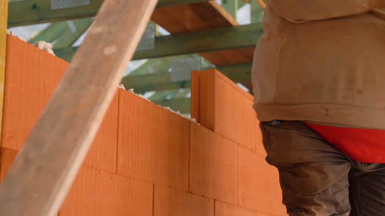 Worker places clay brick onto foam insulation inside unfinished house wall