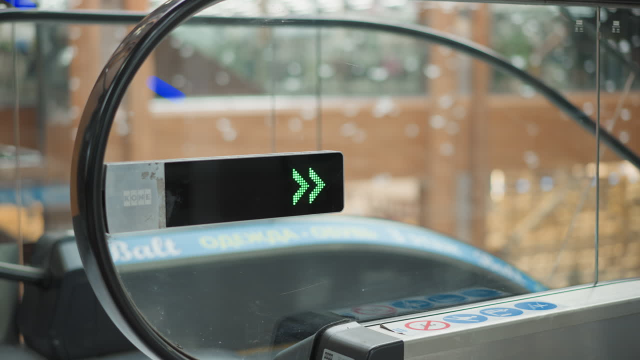 side view of glass mounted green led arrow pointing down at moving walkway entrance with blurred well lit mall hall and moving elevator softly reflected on glass under ambient lighting